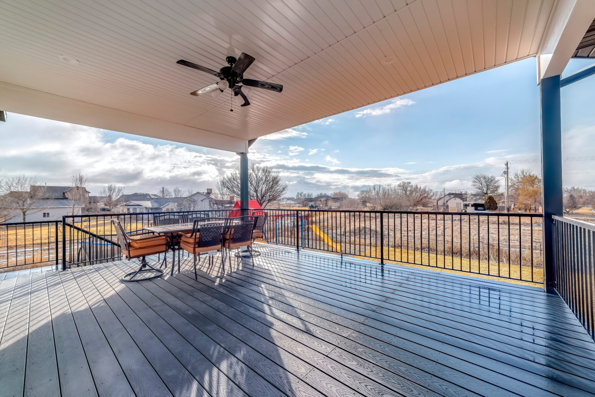 Dining table chairs and ceiling fan with light on wooden deck with metal railing. Yard with playground and neighborhood against cloudy sky can be seen from the home.